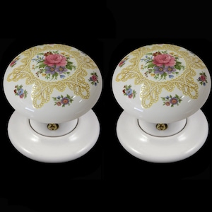 May include: Two white ceramic door knobs with a floral design. The knobs have a gold lace pattern and feature pink, purple, and yellow flowers.