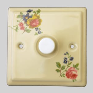 May include: A cream-coloured ceramic light switch plate with a floral design. The switch plate has a single round button with a white centre.