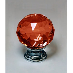 Könnte beinhalten: Ein facettierter Kristall-Schrankknopf mit einer verchromten Basis. Der Kristall ist in einer leuchtenden orangen Farbe.