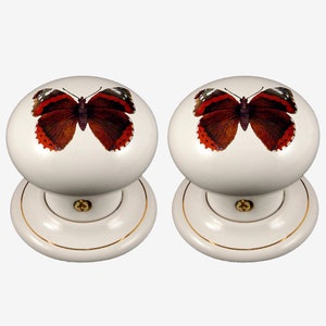 May include: Pair of white ceramic door knobs with a gold trim. Each knob features a red admiral butterfly design.