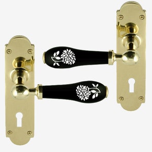 May include: Two gold door handles with black ceramic levers featuring white floral designs. The handles have a keyhole and are mounted on a rectangular plate.
