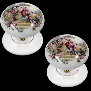 Könnte beinhalten: Zwei weiße Keramik-Türknöpfe mit einem Blumendesign, das eine Frau und ein Kind in einem Garten zeigt.