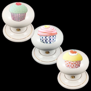 May include: Three white ceramic door knobs with a gold trim. Each knob features a different cupcake design. The first knob has a green cupcake with pink frosting and a red cherry. The second knob has a pink cupcake with yellow frosting and a lemon slice. The third knob has a yellow cupcake with white frosting and a red strawberry.
