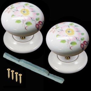 Peut inclure: Deux boutons de porte en céramique blanche avec un motif floral. Les boutons présentent des roses roses avec des centres jaunes et des feuilles vertes. Les boutons sont fixés à des bases en céramique blanche.