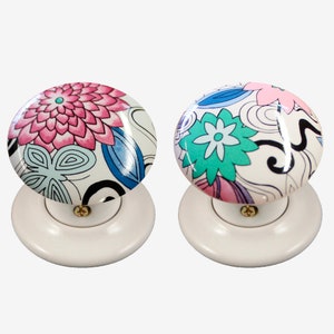 May include: Two white ceramic door knobs with floral designs. One knob features pink and blue flowers, while the other has green and blue flowers.