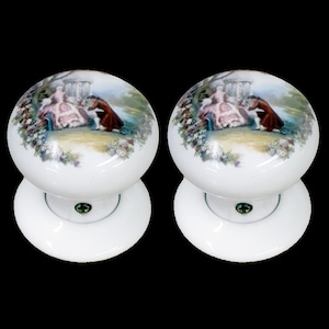 May include: Pair of white ceramic door knobs with a floral design and a painted scene of a couple in a garden.