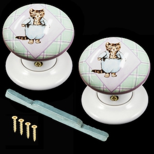 May include: A pair of white ceramic door knobs with a green and purple plaid design and a cartoon cat wearing blue pyjamas. The knobs come with a metal spindle and screws.