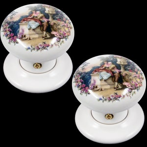 May include: Pair of white ceramic door knobs featuring a floral design and a couple in a garden scene.