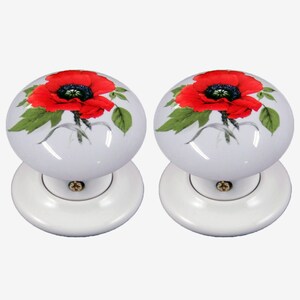 May include: Two white ceramic door knobs with a red poppy flower design. The poppy has a black center and green leaves. The knobs are mounted on white circular backplates with gold-colored screws.