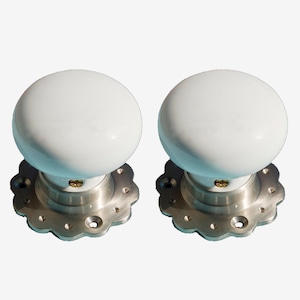 May include: Two white ceramic door knobs with silver metal bases and decorative edges. The knobs are round and have a smooth finish.