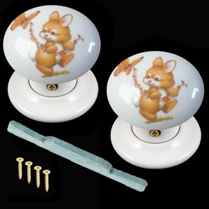 May include: Two white ceramic door knobs featuring a charming bunny rabbit and butterfly design. The knobs are mounted on white bases and include a silver metal spindle and four gold screws.