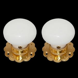 Op de afbeelding: Twee witte keramische deurknoppen met gouden metalen bases en decoratieve gegolfde randen.