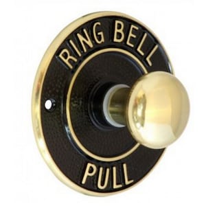 May include: A circular doorbell in black with gold lettering that reads "RING BELL PULL". It features a gold pull knob and a gold trim. The doorbell is designed for pulling.