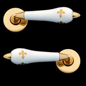 May include: Two white ceramic door handles with gold accents and a fleur-de-lis design. The handles are attached to round gold bases.