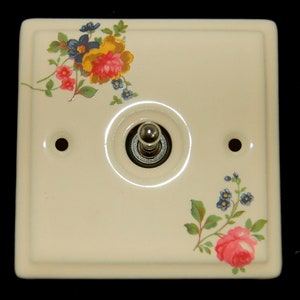 May include: A white ceramic light switch cover with a floral design. The switch cover has a silver toggle switch and two small holes on either side. The floral design features pink, yellow, and blue flowers.