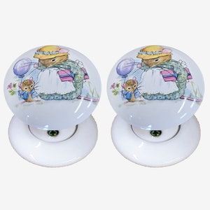 以下が含まれることがあります： 青い背景に、プレゼントと風船を持ったネズミの絵が描かれた白いセラミック製のドアノブが2つ。ネズミは黄色の帽子と青いドレスを着ています。