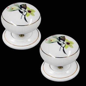 以下が含まれることがあります： 金色の縁取りと花柄の白いセラミック製のドアノブが2つ。花柄は緑の葉、白い花、赤い実が特徴です。