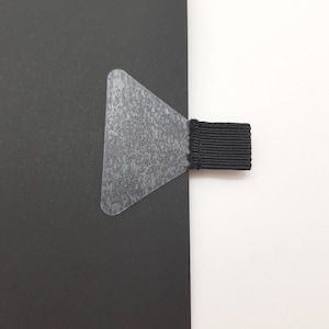 Op de afbeelding: Een zwart notitieboek met een zwart lint-bladwijzer en een doorzichtige hoekbeschermer van plastic.