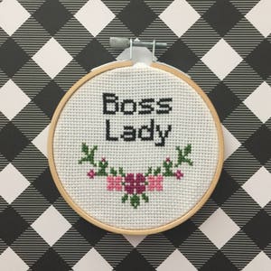 Może przedstawiać: Biały tamborek do haftu z napisem "Boss Lady" czarnymi literami i wzorem kwiatowym z różowymi kwiatami i zielonymi liśćmi.