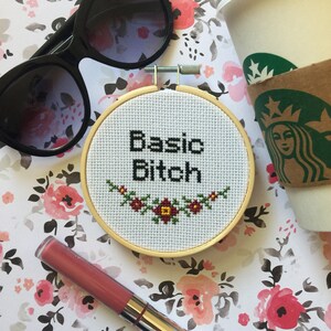 Puede incluir: Un aro de bordado blanco con la frase "Basic Bitch" bordada en hilo negro. El aro está rodeado de un patrón floral sobre un fondo blanco.