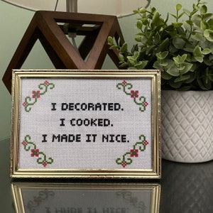 May include: A framed cross-stitch with a gold frame and white background. The cross-stitch reads "I decorated. I cooked. I made it nice."