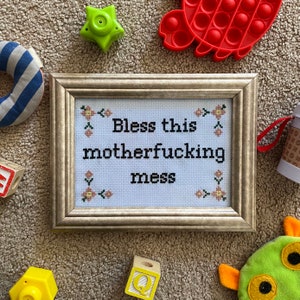 May include: A framed cross-stitch with the text "Bless this motherfucking mess" in black lettering on a light blue background. The cross-stitch is surrounded by a brown frame and is surrounded by various toys, including a pop-it toy, blocks, and a stuffed animal.