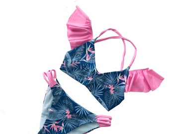Peut inclure: Un maillot de bain deux pièces avec un motif floral tropical. Le haut présente un imprimé floral bleu et blanc avec des accents roses et des volants roses. Le bas est bleu et blanc avec des bretelles roses.