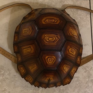 May include: A brown leather backpack shaped like a turtle shell. The shell is made of multiple pieces of leather sewn together to create a realistic turtle shell design. The backpack has two adjustable straps.