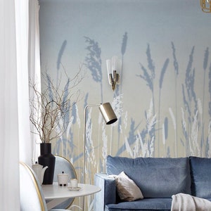 May include: A living room interior with a blue velvet sofa, two light blue chairs with gold accents, a white round table, and a floor lamp. The wall features a light blue wallpaper with a white silhouette of tall grass.