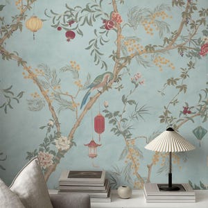 Könnte beinhalten: Hellblaue Tapete mit detailliertem Chinoiserie-Design. Das Muster zeigt Äste, Vögel, Laternen und Blumen in Braun-, Grün-, Rot- und Gelbtönen. Im Vordergrund ein weißer Tisch mit Lampe, Büchern und Vase.