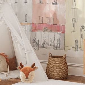 Op de afbeelding: Een witte tipi-tent met bruine kwastjes en pompoenen, een bruin en oranje pluche herten speelgoed, een bruin en wit pluche paard speelgoed, een bruine rieten mand en een witte muur met een handgetekende illustratie van een straat in de stad met een winkel die "Flowers" zegt.
