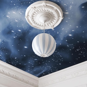 May include: A spherical pendant light with vertical light blue and white stripes suspended from a white ornate ceiling medallion. The ceiling is painted with a dark blue night sky mural featuring white stars and soft, cloudy textures.