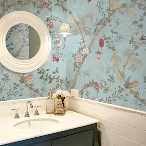 May include: A bathroom vanity with a white marble countertop, a chrome faucet, and two glass bottles. Above the vanity is a round white mirror and a wall sconce. The walls feature light blue wallpaper with a pattern of branches, flowers, and birds.