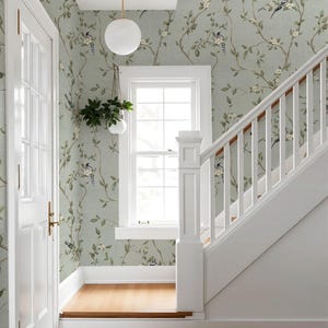 May include: Light sage green wallpaper featuring a repeating pattern of brown branches, green leaves, small white flowers, and blue birds. The wallpaper covers the walls of a staircase landing with a white window frame and a white staircase railing.