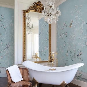 Puede incluir: Una ba&ntilde;era blanca con patas doradas y grifer&iacute;a dorada, situada frente a un gran espejo con marco dorado. Una silla de madera con asiento y respaldo de rejilla sostiene una toalla blanca, sobre un papel pintado azul claro con motivos de aves.