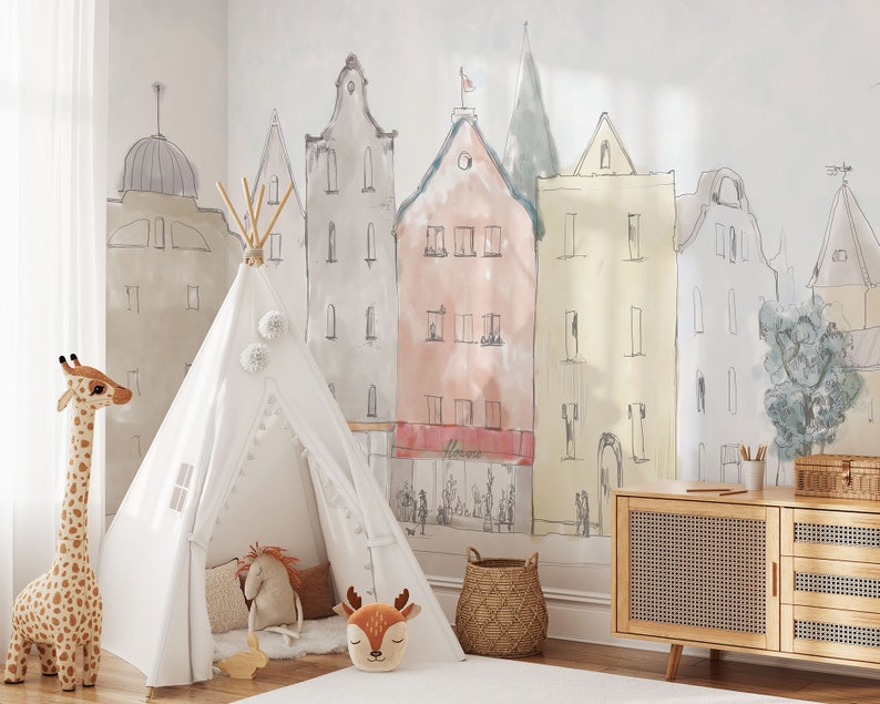 Op de afbeelding: Een witte tipi-tent met een witte pompon-bies en een bruin giraffenknuffeldier. De tipi staat in een kamer met een muurschildering van een stadsbeeld dat op de muur is geschilderd. Er is een houten dressoir met een geweven rotanfront en een rieten mand in de kamer.