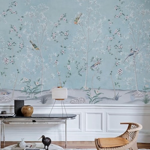 Puede incluir: Un papel pintado azul claro con un mural de estilo pintado a mano de &aacute;rboles delgados, flores en flor y p&aacute;jaros peque&ntilde;os. El dise&ntilde;o incluye una l&iacute;nea de suelo gris suave con rocas y follaje, creando una escena natural serena en la pared.