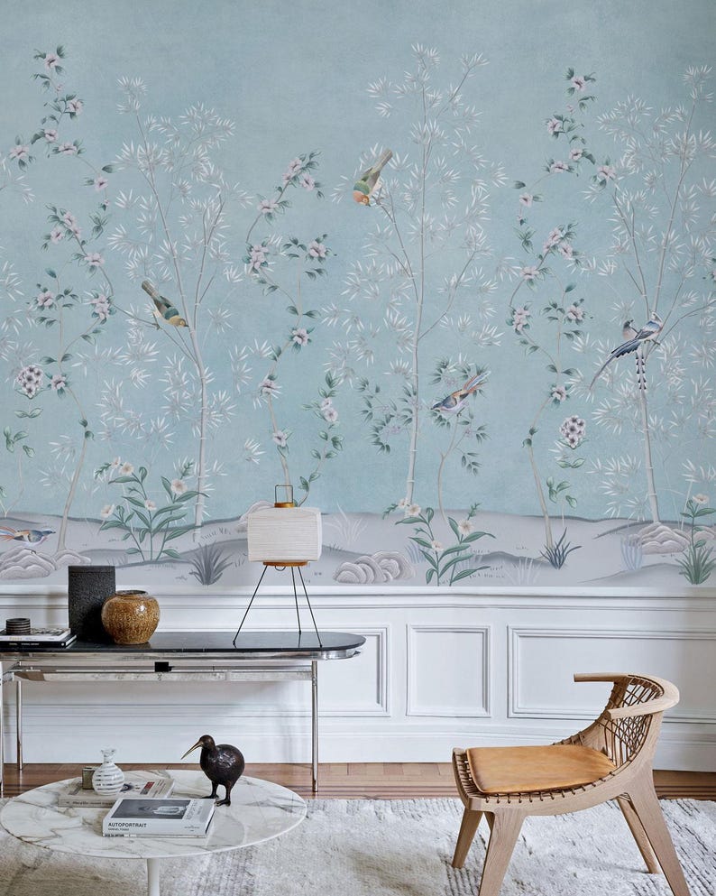 Puede incluir: Un papel pintado azul claro con un mural de estilo pintado a mano de &aacute;rboles delgados, flores en flor y p&aacute;jaros peque&ntilde;os. El dise&ntilde;o incluye una l&iacute;nea de suelo gris suave con rocas y follaje, creando una escena natural serena en la pared.