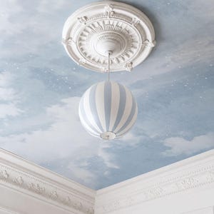 May include: A spherical pendant light with vertical light blue and white stripes suspended from a white ornate ceiling medallion. The ceiling is painted with a blue sky mural featuring white clouds and small white stars, framed by white crown molding.