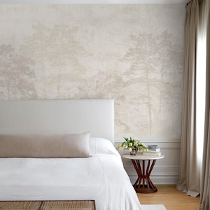 May include: A bedroom featuring a wall covered in wallpaper with a muted, monochromatic forest landscape print in shades of beige and cream. The room includes a white upholstered headboard, a white bedspread, and a small round side table.