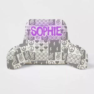 May include: A gray and white patterned reading pillow with the name "SOPHIE" in purple text. The pillow features peace signs, lightning bolts, smiley faces, and hearts.