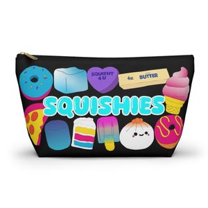 May include: Black zippered pouch featuring colorful illustrations of squishies, including a donut, ice cream cone, pizza slice, and a heart-shaped candy with the text "Squishy 4 U". The word "SQUISHIES" is written in large, colorful letters.