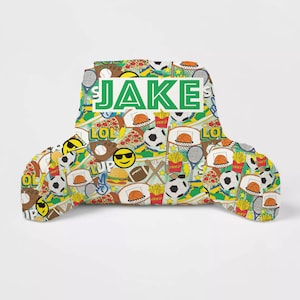 May include: A green and white reading pillow with a colorful sports and food pattern. The pillow has a large green rectangle with the name "JAKE" in white letters. The pillow is perfect for reading or relaxing.