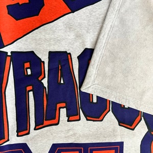 K&ouml;nnte beinhalten: Grauer Sweatshirt mit den Worten "Syracuse" und "Orange" in blauer und orangefarbener Schrift.