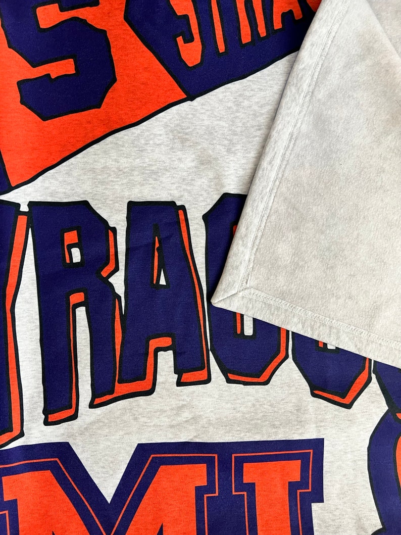 K&ouml;nnte beinhalten: Grauer Sweatshirt mit den Worten "Syracuse" und "Orange" in blauer und orangefarbener Schrift.