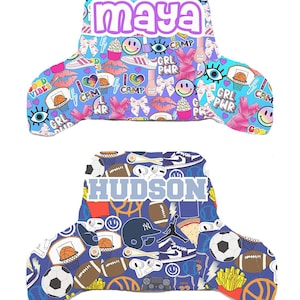 May include: Two personalized car seat headrest covers. The top one is light blue with the name "maya" in purple, and features a pattern of basketballs, bows, and smiley faces. The bottom one is blue with the name "HUDSON" and sports-themed graphics.