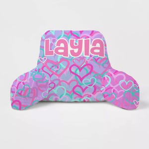 May include: A pink and purple reading pillow with a heart pattern and the name "Layla" printed on the front.