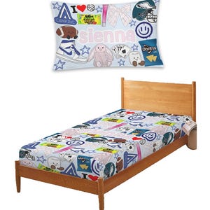 May include: A twin-sized bed with a light wood frame and a patterned bedspread. The bedspread features various colorful graphics, including a football, a bunny, and the name "Sienna". A matching pillow with similar graphics is on the bed.