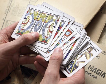 May include: A hand holds a deck of tarot cards, with the top card showing a woman with wings and a crown. The cards are on a table with a book titled "MAGIA PSYCHOLOGII" visible in the background.
