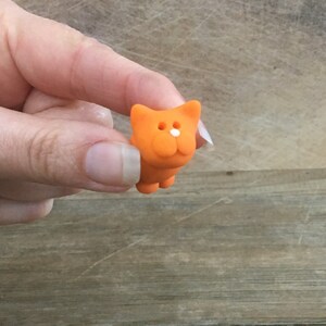 May include: A small, orange cat figurine with a white nose and two white eyes. The cat is held in a hand against a wooden background. The figurine has a simple, cartoonish design.